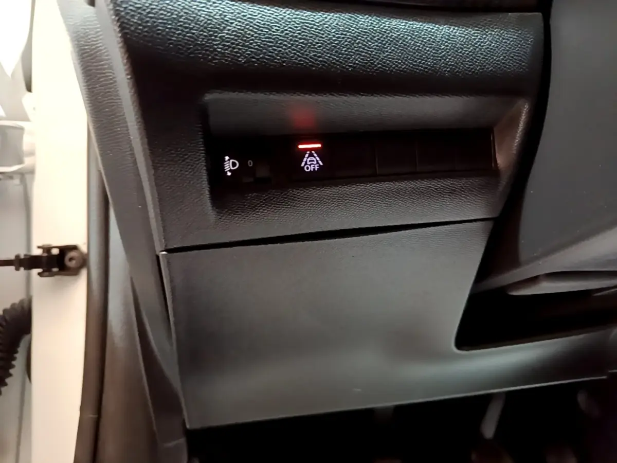 Gros plan sur les commandes sous le tableau de bord côté gauche d'une Peugeot 208 blanche, avec témoin d'alerte allumé.