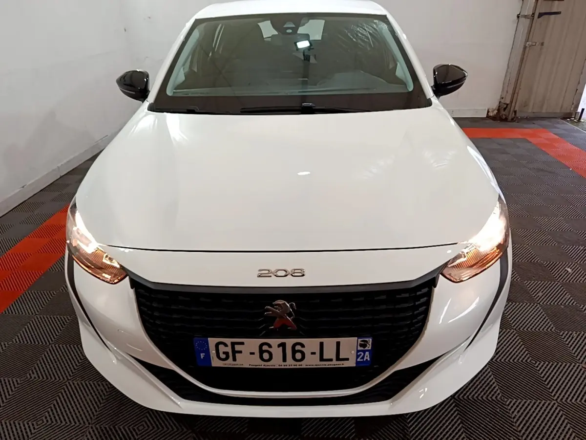 Vue avant rapprochée d’une Peugeot 208 blanche avec phares allumés dans un garage.