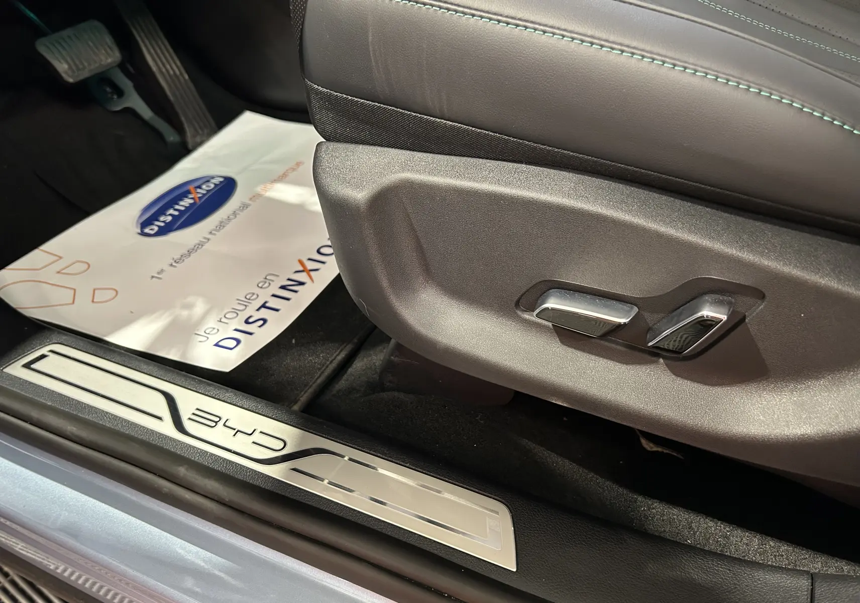 Gros plan sur la commande de réglage du siège en cuir noir et la plaque de seuil BYD côté conducteur, intérieur lumineux.