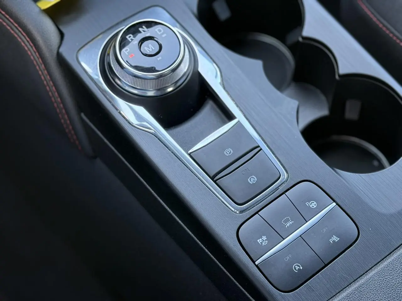 Gros plan sur la console centrale du Ford Kuga 2024 rouge Lucifer, avec sélecteur rotatif de boîte auto et commandes d’assistance.