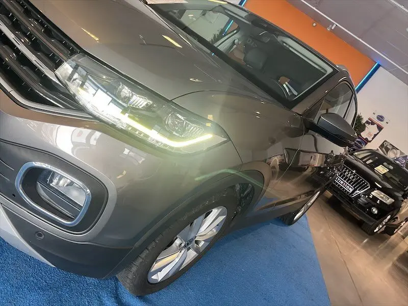 Vue 3/4 avant droit d'un Volkswagen T-Cross gris Limestone métallisé avec phares LED allumés en showroom.