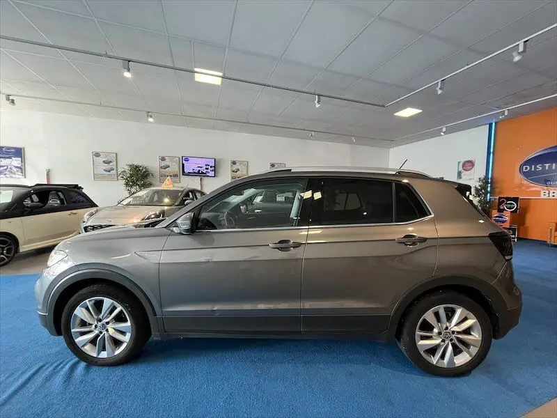 Profil côté gauche d'un Volkswagen T-Cross gris Limestone métallisé exposé en showroom sur sol bleu.