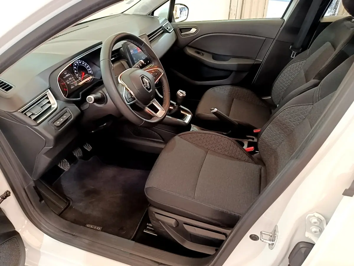 Vue intérieure côté conducteur d'une Renault Clio Business blanche, montrant sièges noirs et tableau de bord avec volant multifonctions.