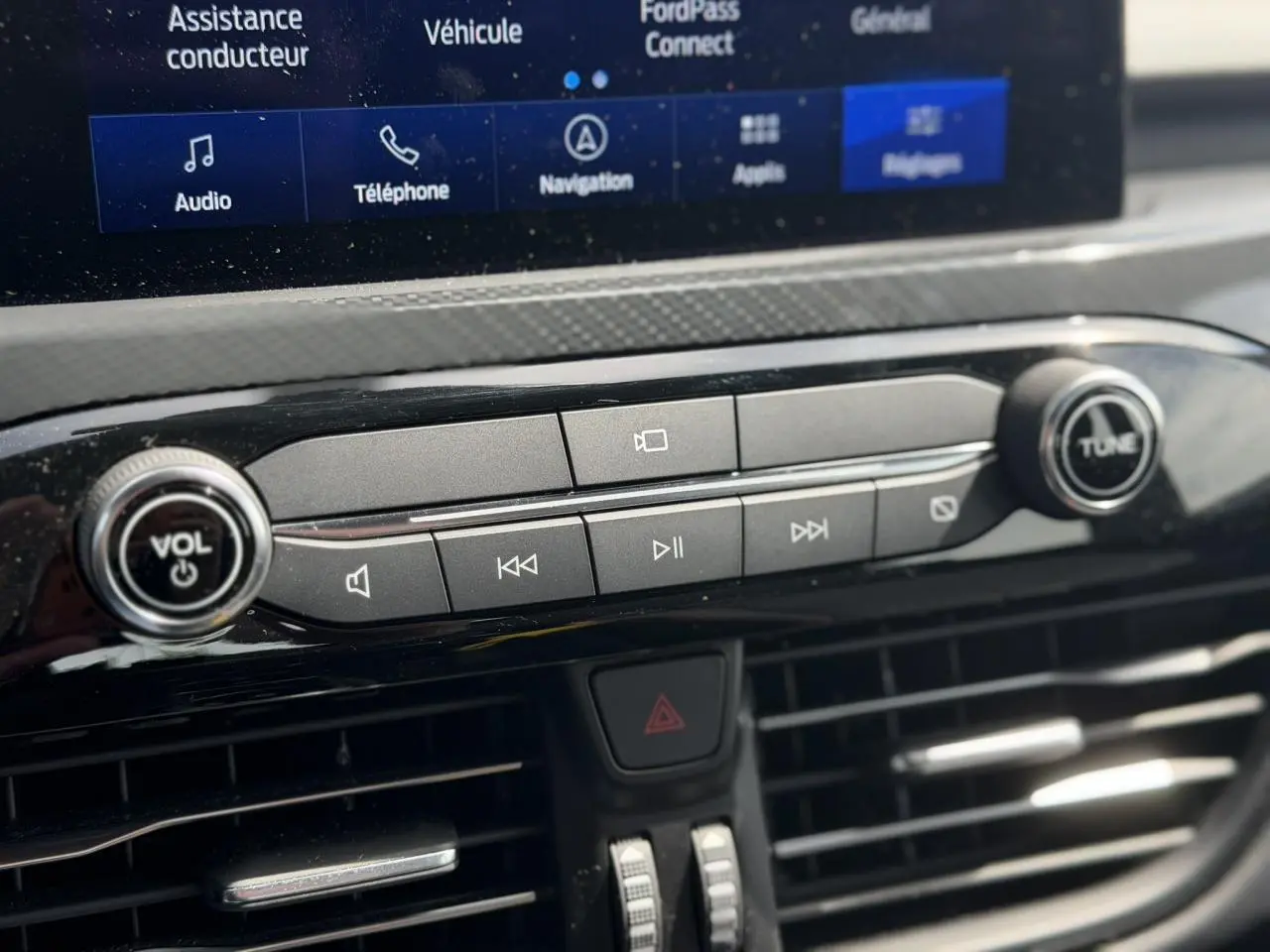Gros plan sur la console centrale du Ford Kuga 2024, boutons de contrôle audio et écran tactile visible en arrière-plan.