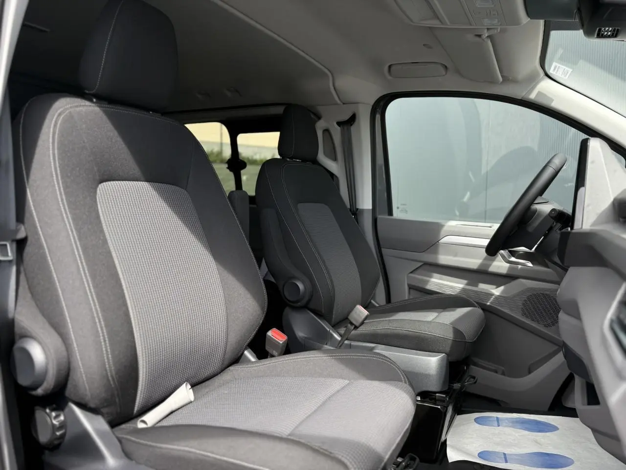 Vue intérieure côté conducteur du Volkswagen Transporter T7 ProCab 2026, sièges tissu gris à rayures et volant multifonction.