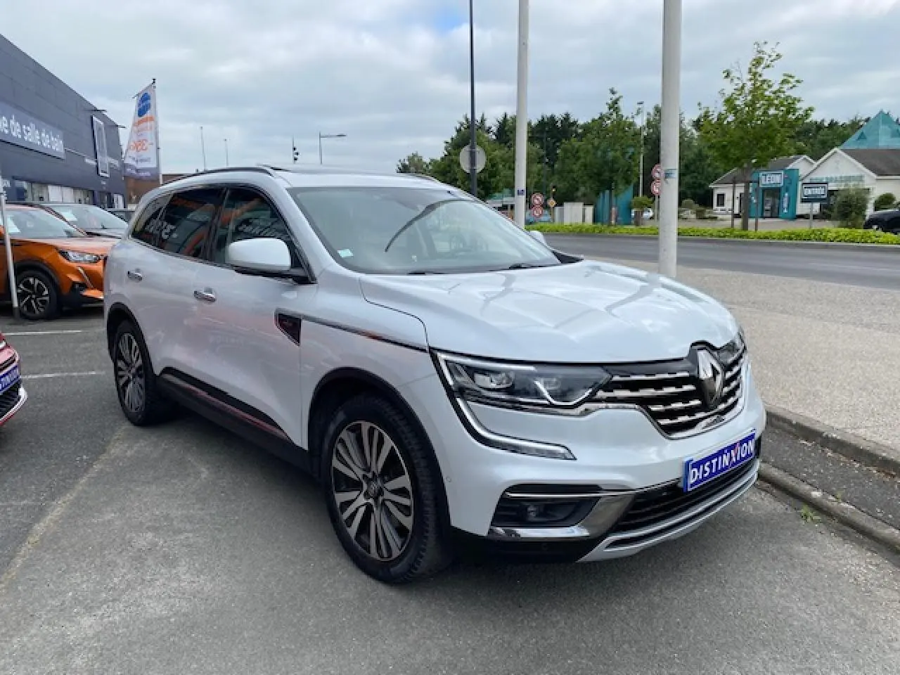 Renault Koleos blanc en 3/4 avant droit, avec calandre chromée et jantes alliage distinctives, stationné en extérieur.