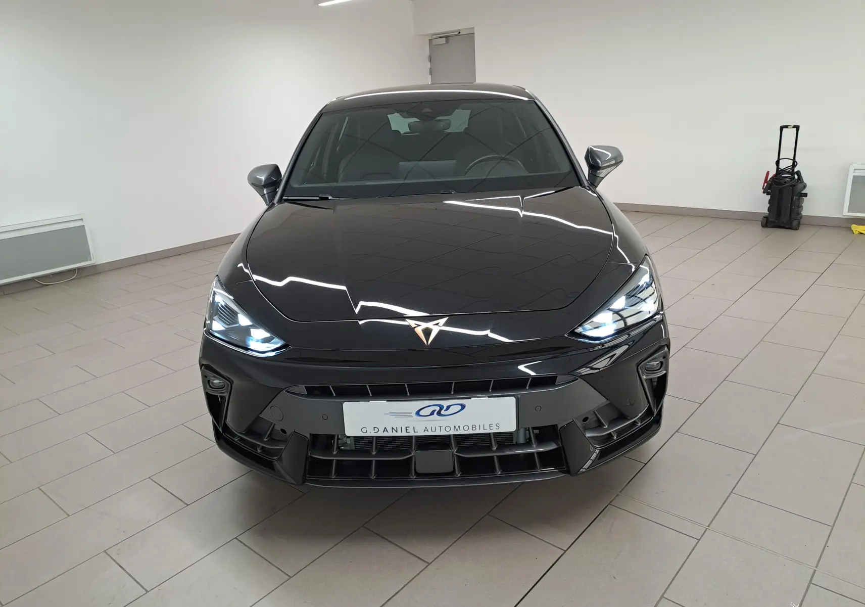 Vue avant d'une CUPRA Leon Ph.2 2025 en Noir Minuit avec phares allumés et logo cuivré distinctif.