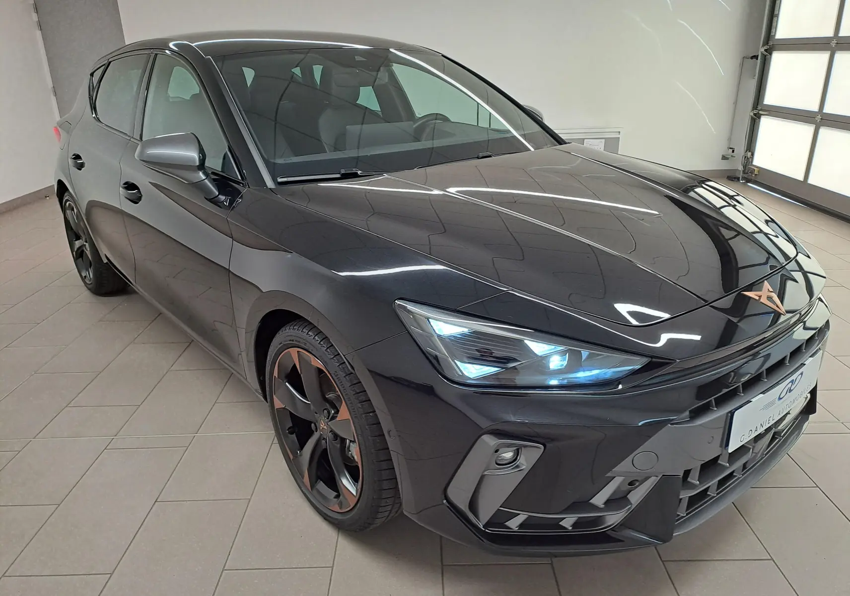 Vue 3/4 avant d'une CUPRA Leon 2025 Noir Minuit avec jantes cuivrées et phares LED allumés en intérieur.