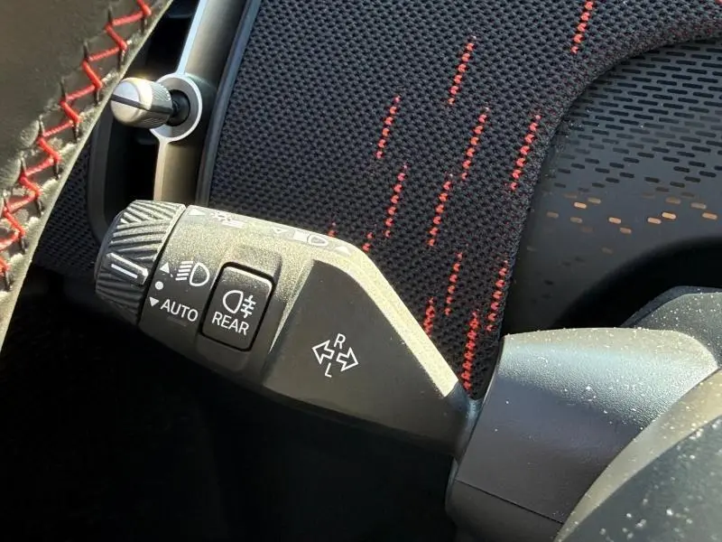 Gros plan sur la commande des feux et clignotants du MINI Countryman JCW, avec tissu noir et surpiqûres rouges en arrière-plan.