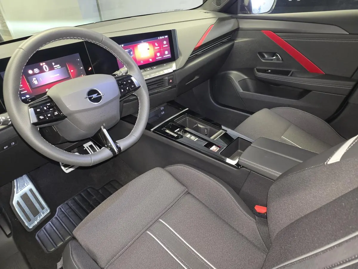Vue intérieure côté conducteur de l’Opel Astra Hybrid 2024 noire, avec tableau de bord digital et touches rouges décoratives.