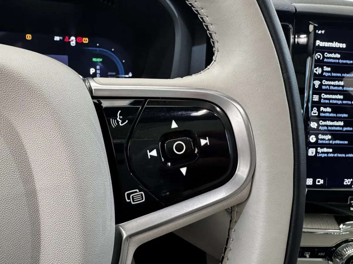 Gros plan sur les commandes du volant cuir beige du Volvo XC60 bleu, affichage numérique et écran tactile visibles.