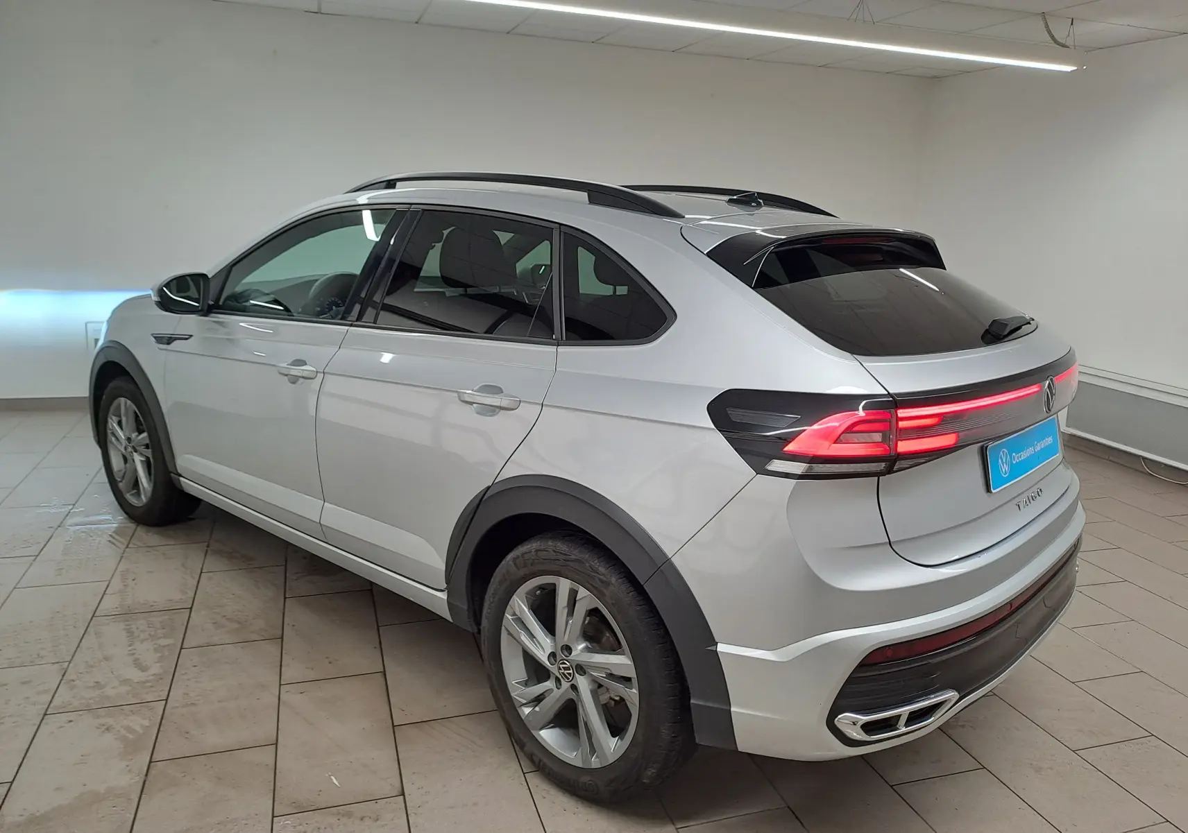 Volkswagen Taigo 2024 R-Line en reflet d'argent, vue 3/4 arrière droit avec feux arrière allumés en intérieur.