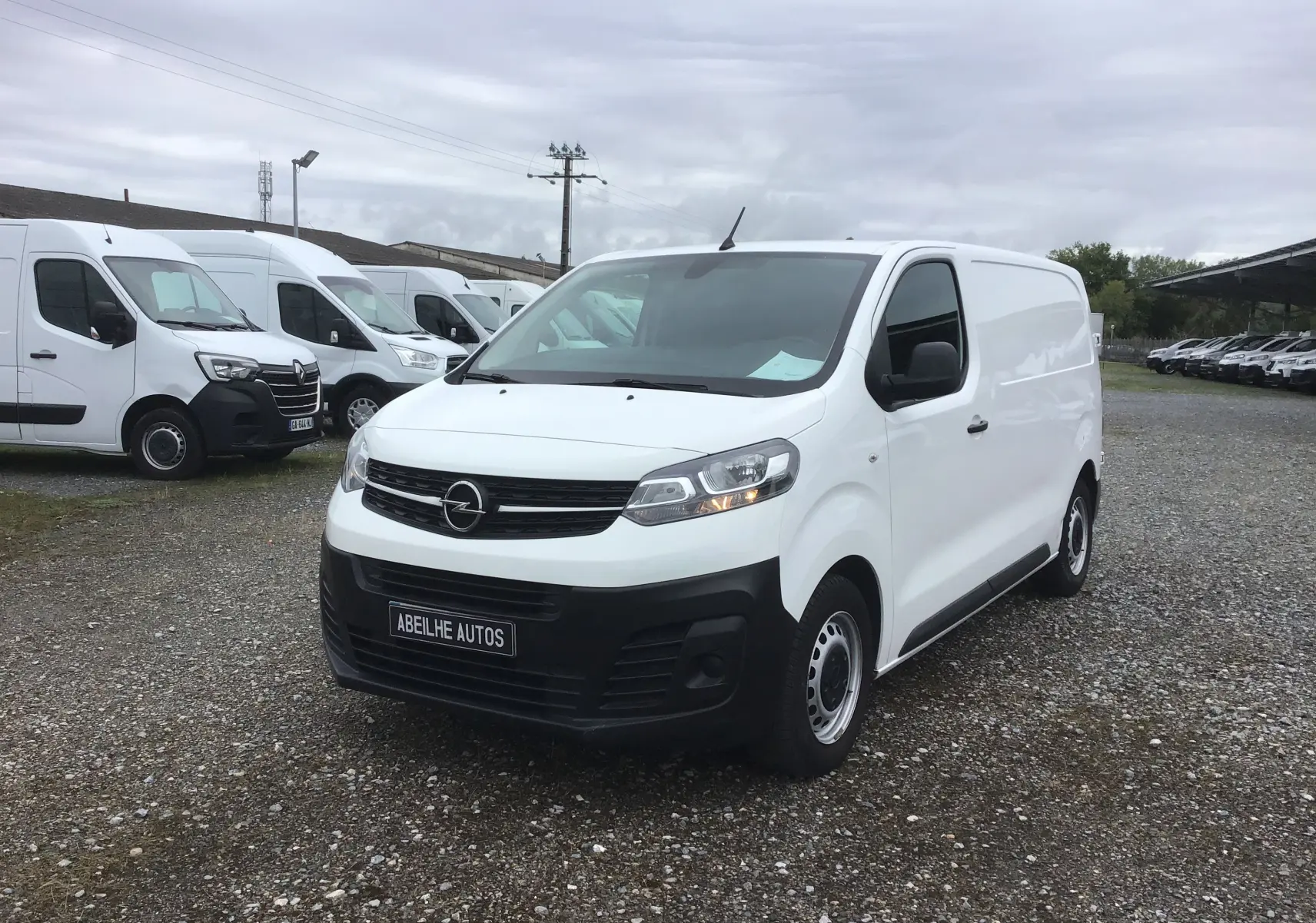Opel Vivaro blanc en 3/4 avant droit sur un parking avec plusieurs fourgons blancs en arrière-plan.