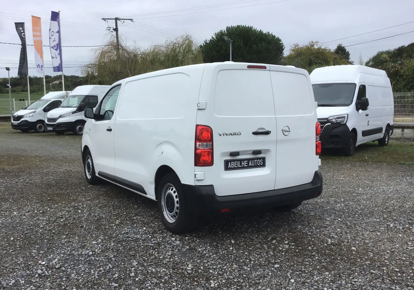 Vue 3/4 arrière droite d’un utilitaire Opel Vivaro blanc stationné sur un sol gravillonné avec d’autres fourgons en arrière-plan.