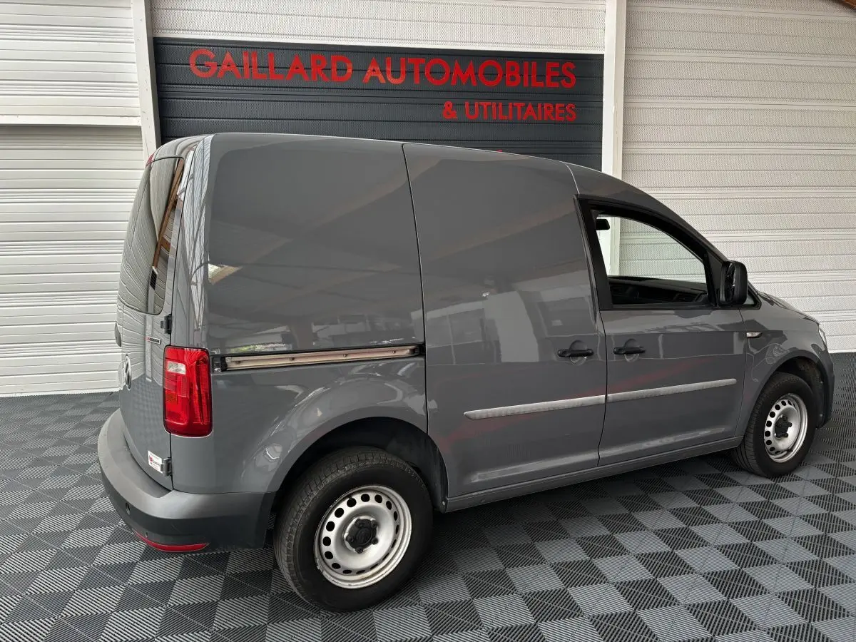 Vue 3/4 arrière côté gauche d'un utilitaire Volkswagen Caddy gris foncé stationné en intérieur sur sol damier.