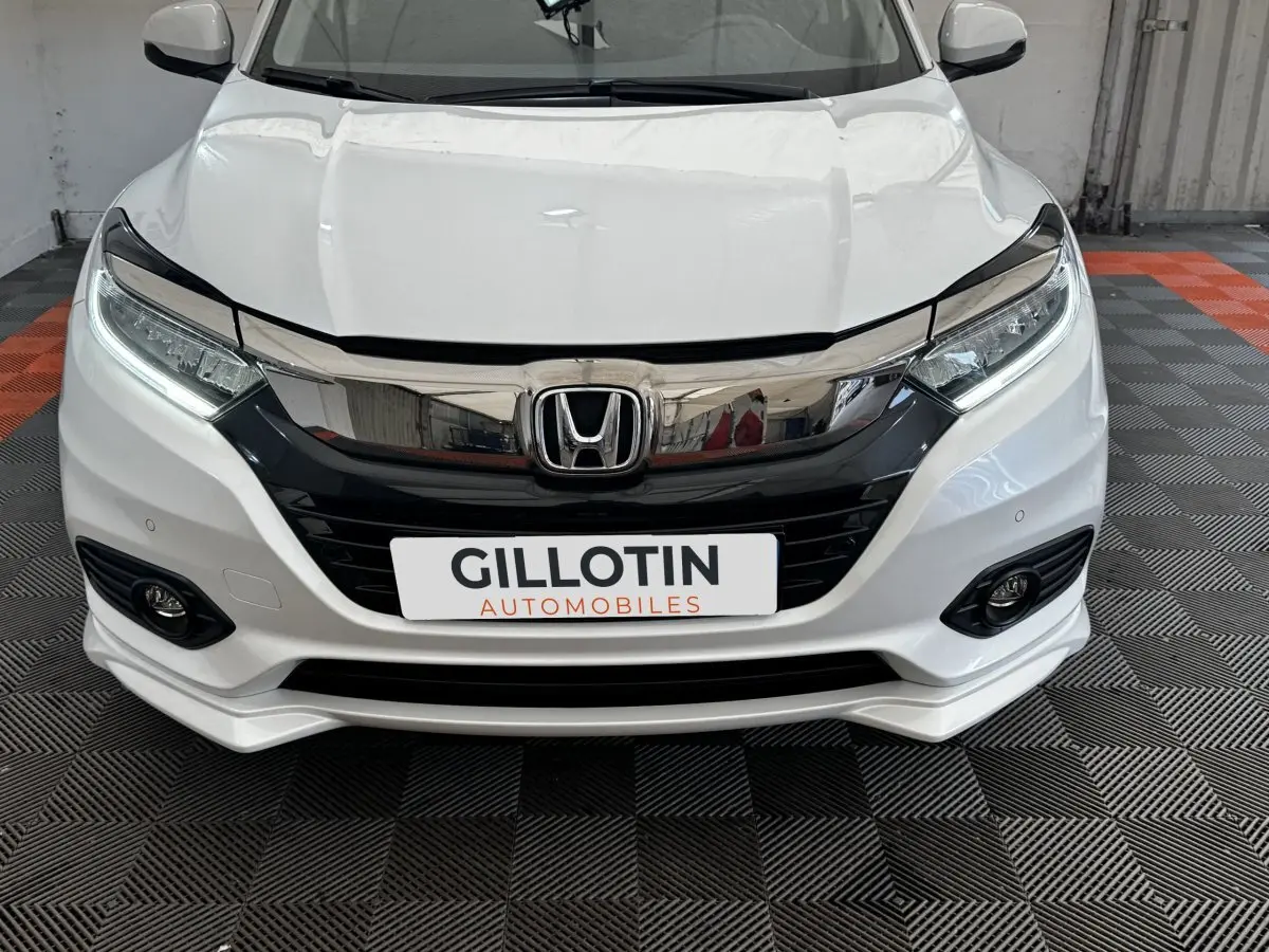 Vue avant rapprochée d’un Honda HR-V blanc avec calandre noire brillante et feux LED, sur sol damier gris et orange.