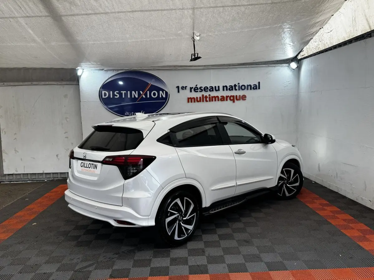 Honda HR-V blanc en vue 3/4 arrière droit, avec jantes noires et toit spoiler dans un showroom.
