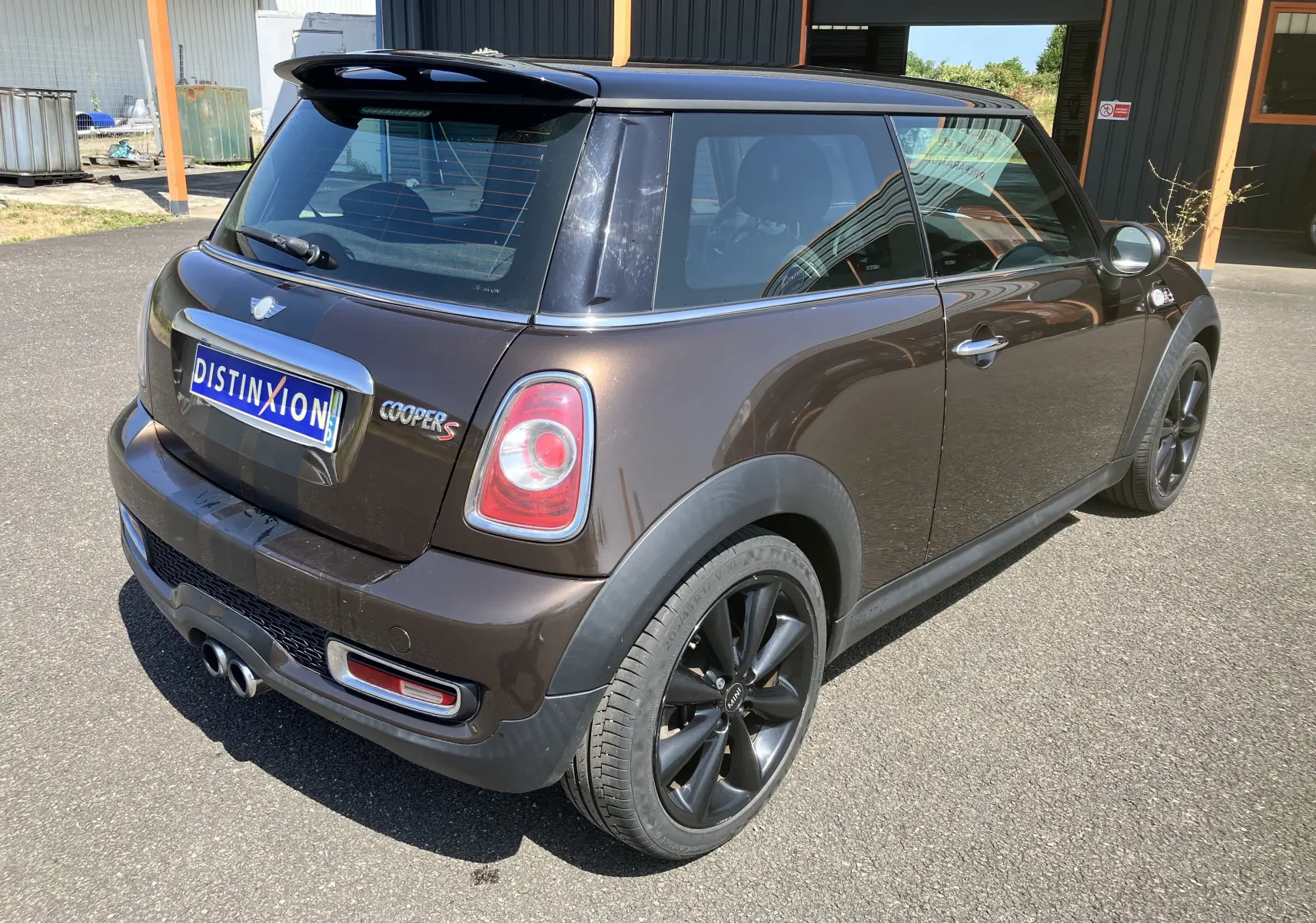 Vue 3/4 arrière droite d'une MINI Cooper S R56 marron avec toit noir et jantes noires sur parking extérieur.
