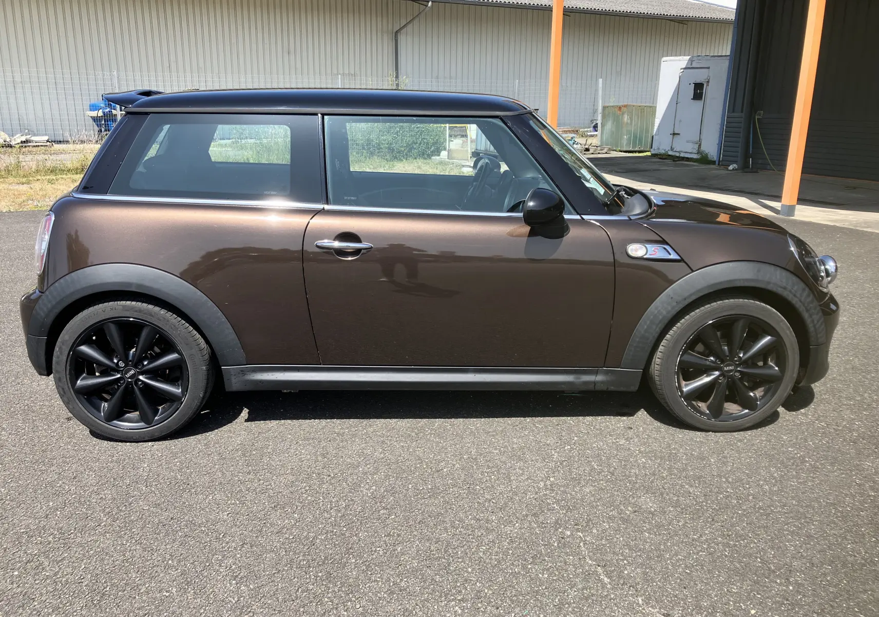 Profil droit d'une MINI Cooper S R56 marron avec toit noir et jantes noires, garée sur un sol goudronné en extérieur.