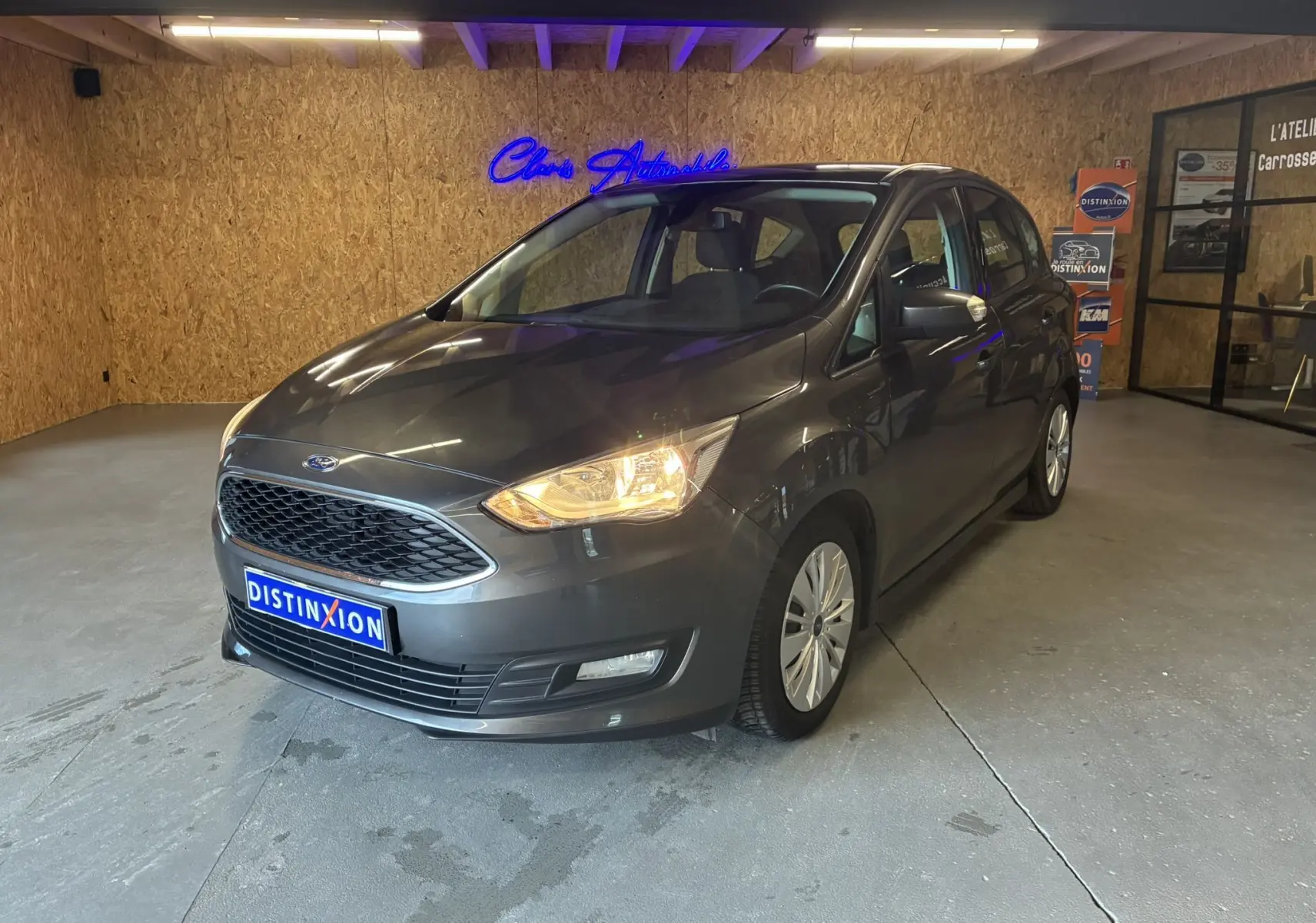 Ford C-MAX gris 3/4 avant droit en intérieur avec phares allumés et calandre hexagonale distinctive.
