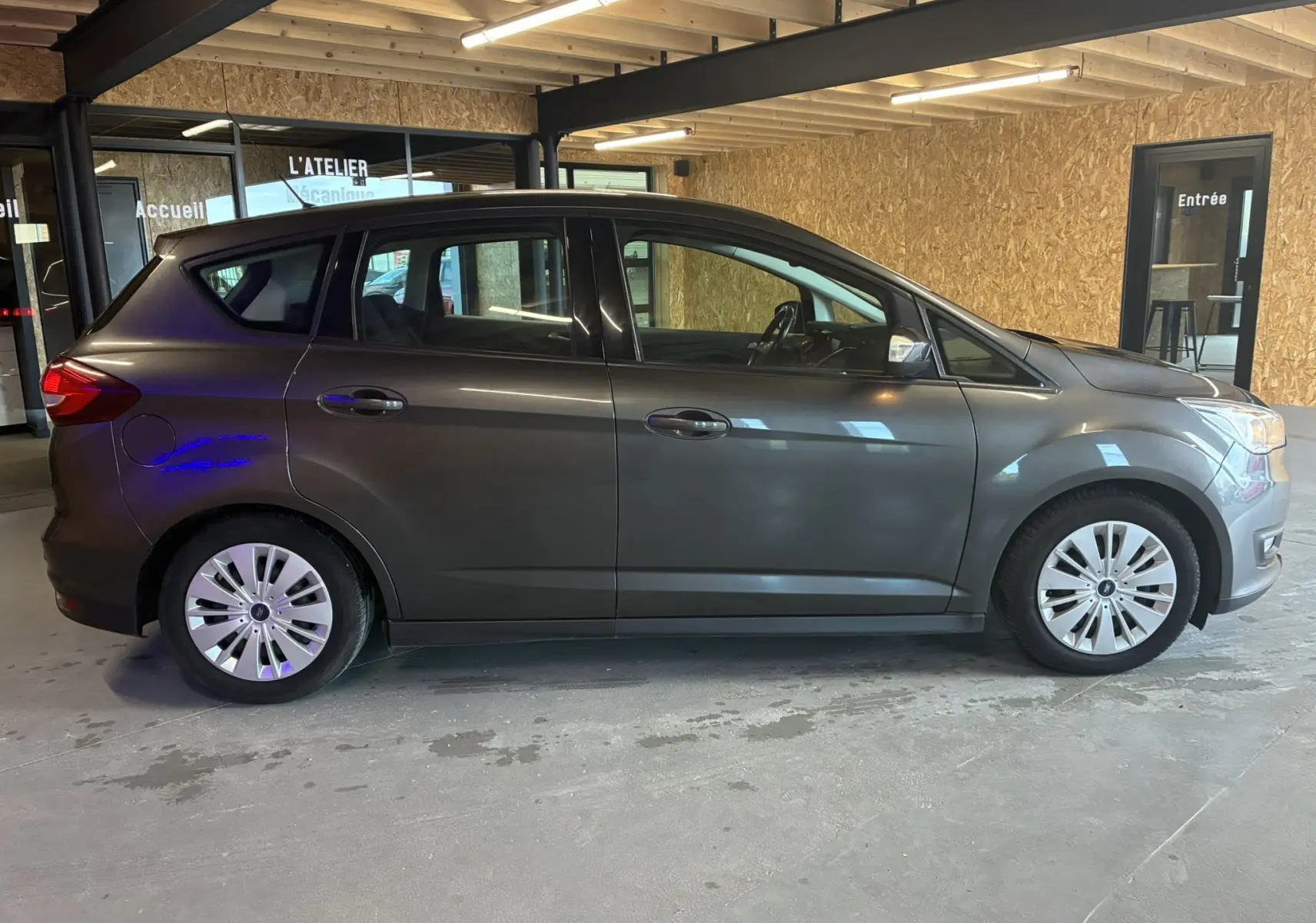 Profil côté gauche d'un Ford C-MAX gris 2015 garé en intérieur, avec jantes argentées et cinq portes visibles.