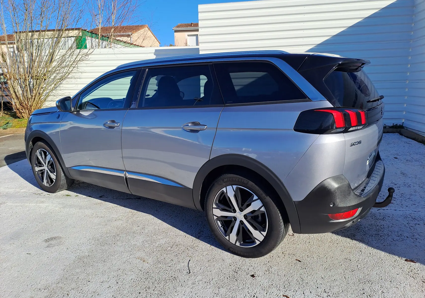 Vue 3/4 arrière droite d’un Peugeot 5008 gris 2019 avec barres de toit et attelage visible.
