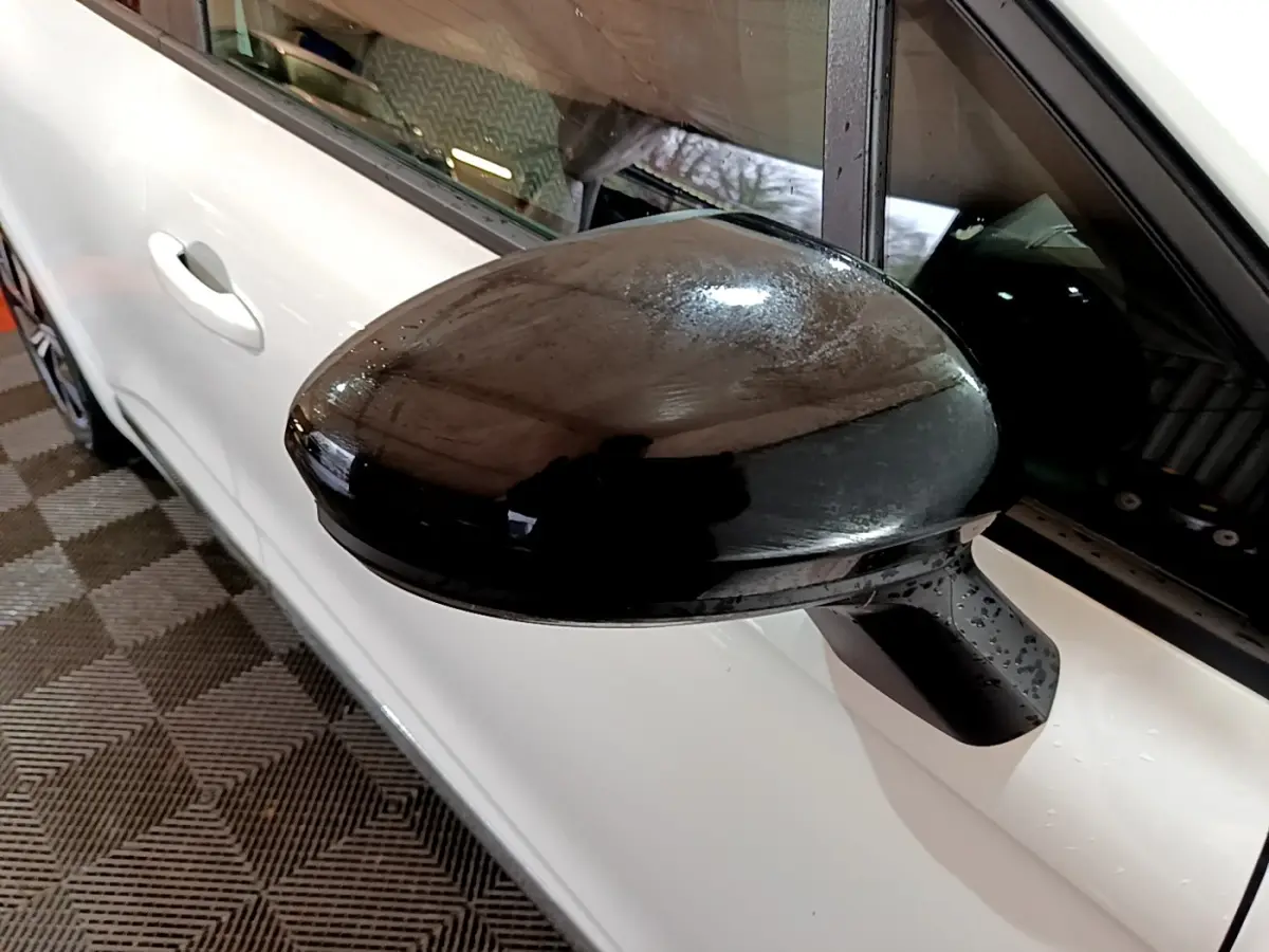 Rétroviseur noir en gros plan sur côté gauche d’une Renault Clio blanche vue de profil.