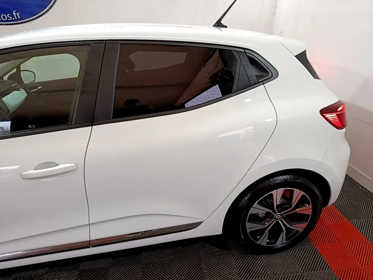 Vue latérale droite d'une Renault Clio blanche 2023 avec jantes alliage et vitres teintées, stationnée en intérieur.