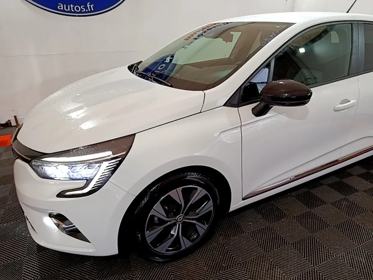 Vue 3/4 avant droit d'une Renault Clio Business blanche avec phares LED allumés et rétroviseurs noirs brillants.