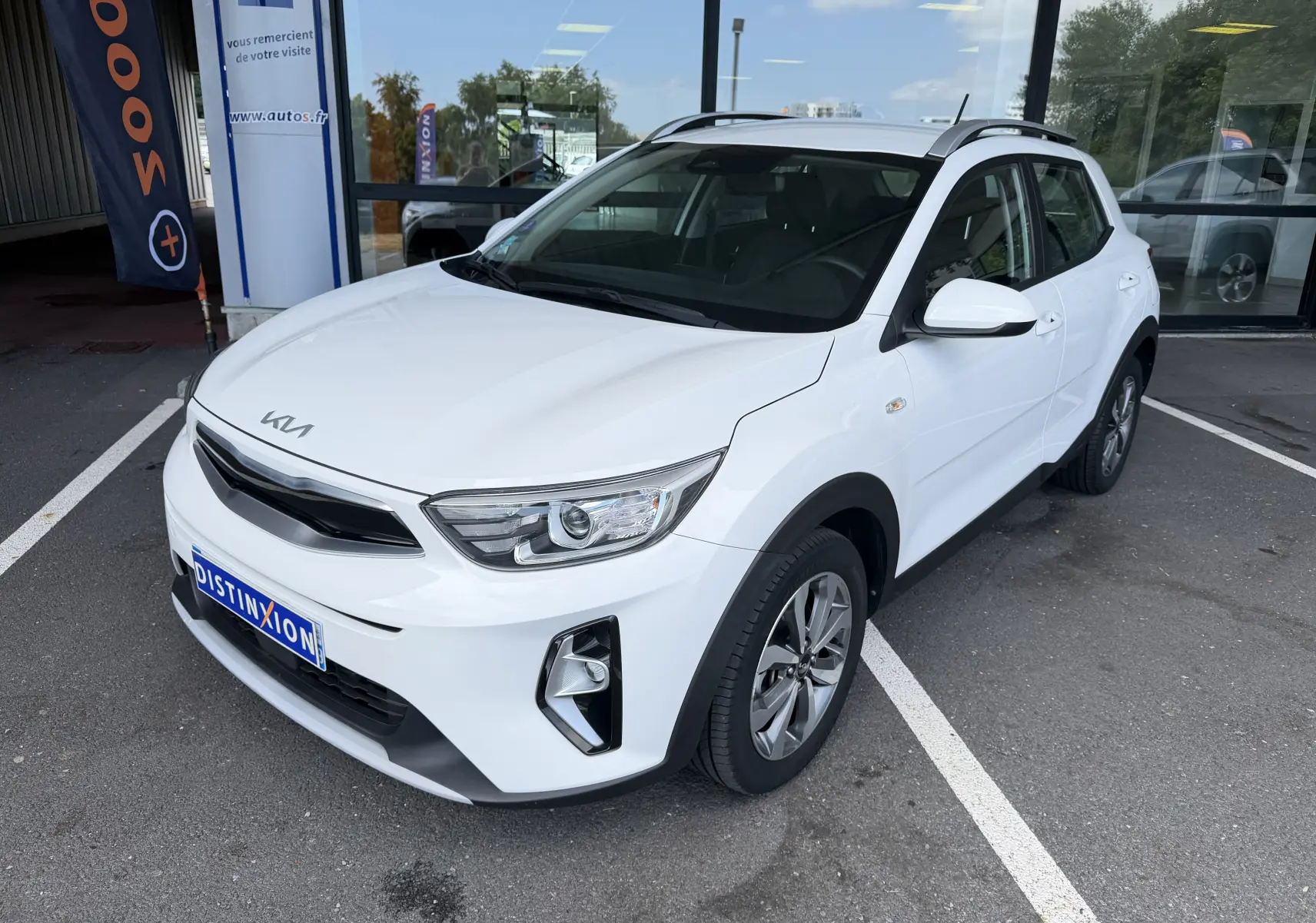 KIA Stonic blanc vue 3/4 avant droit, avec calandre noire et jantes alliage bicolores, stationné devant un bâtiment.