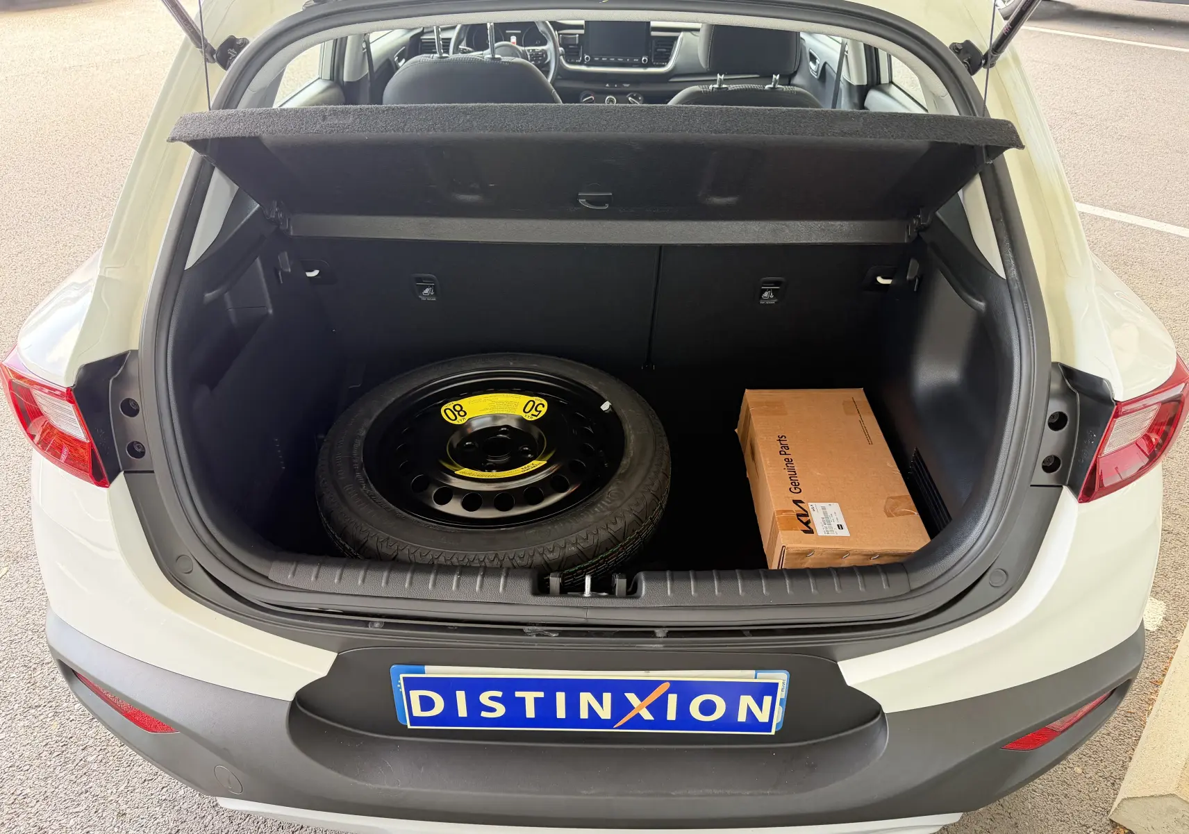 Coffre ouvert d’un KIA Stonic blanc vu de l’arrière, montrant une roue de secours et une boîte en carton à l’intérieur.