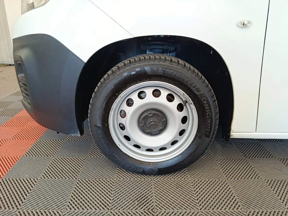 Gros plan sur la roue avant gauche en jante acier du Citroën Berlingo blanc, avec partie basse du pare-chocs noir visible.