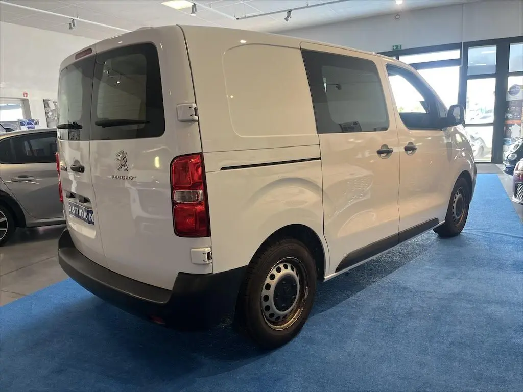 Vue 3/4 arrière droite d'un Peugeot Expert Fourgon blanc stationné en intérieur avec vitres teintées et jantes acier.