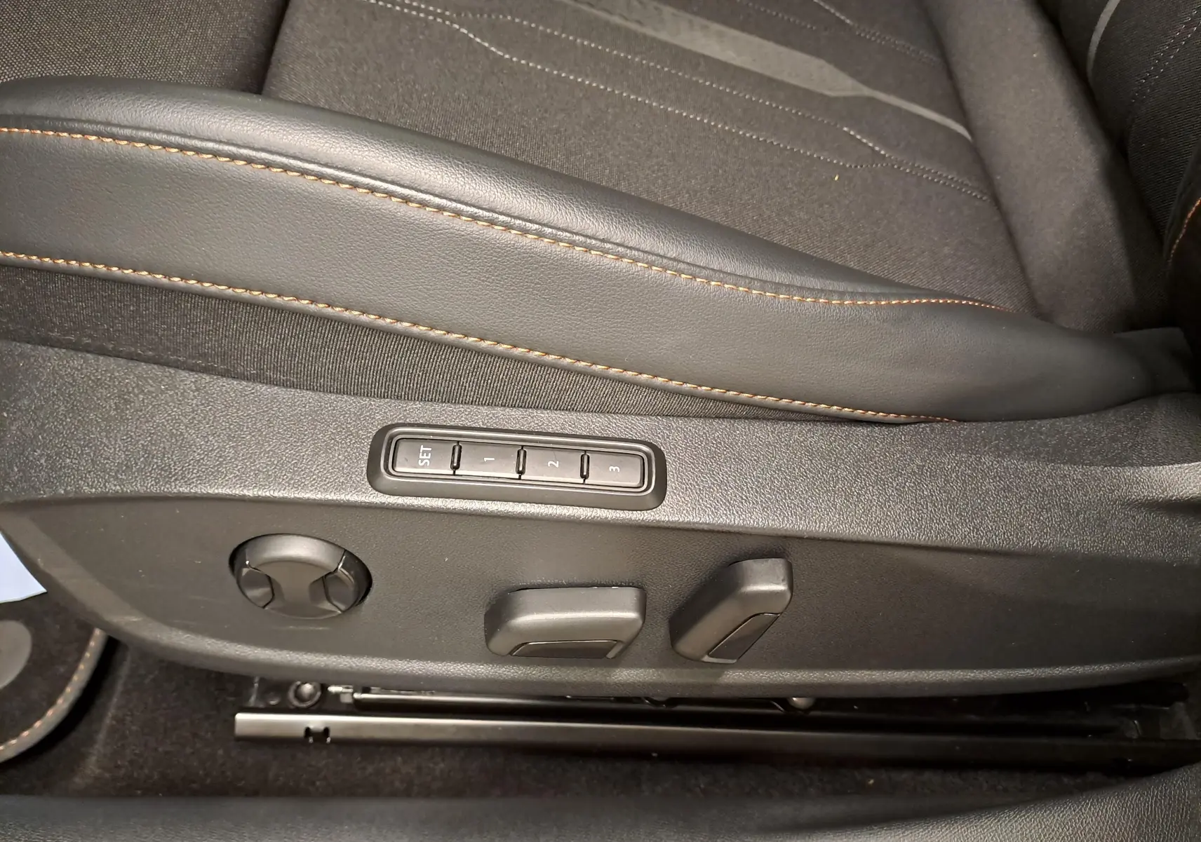 Détail des commandes de réglage électrique du siège conducteur avec surpiqûres orange sur sellerie noire.