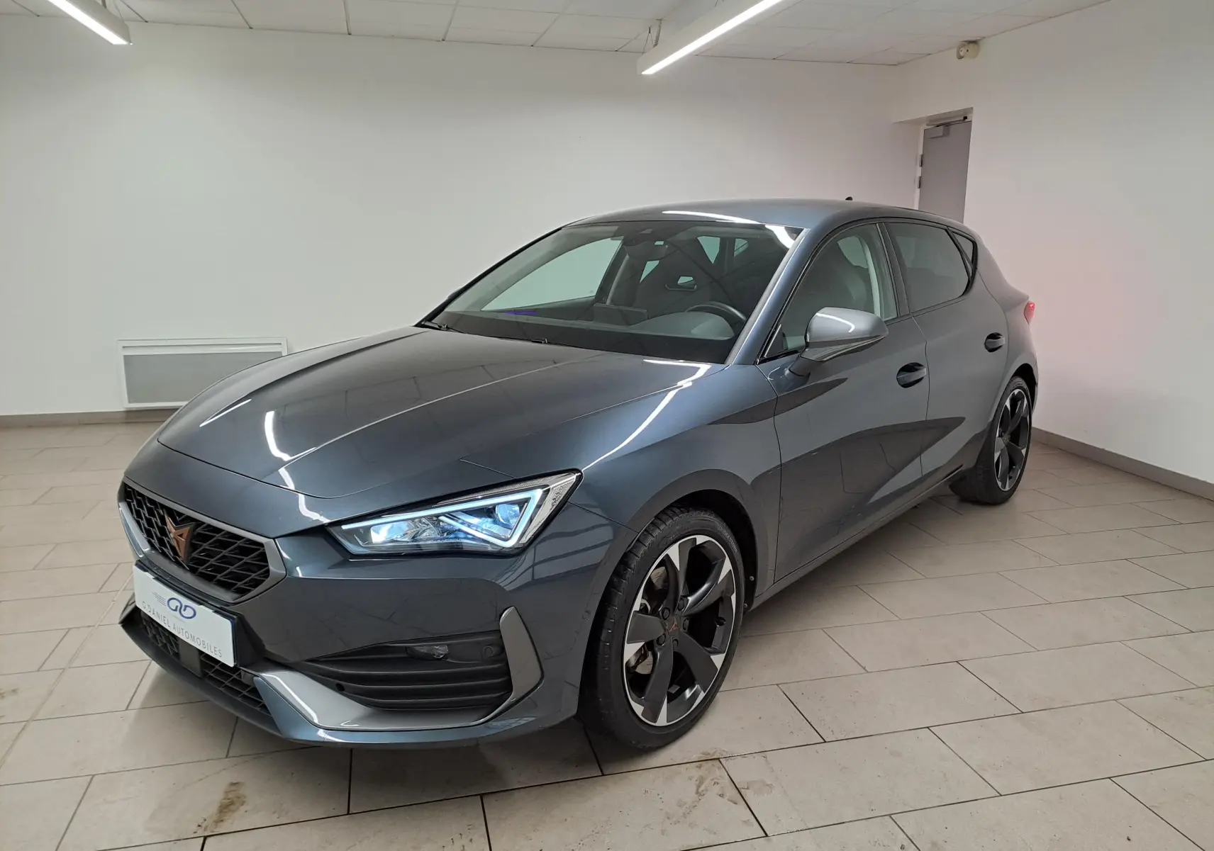 Vue 3/4 avant droite d'une CUPRA Leon 2023 gris magnétique avec jantes noires et phares LED allumés dans un intérieur neutre.