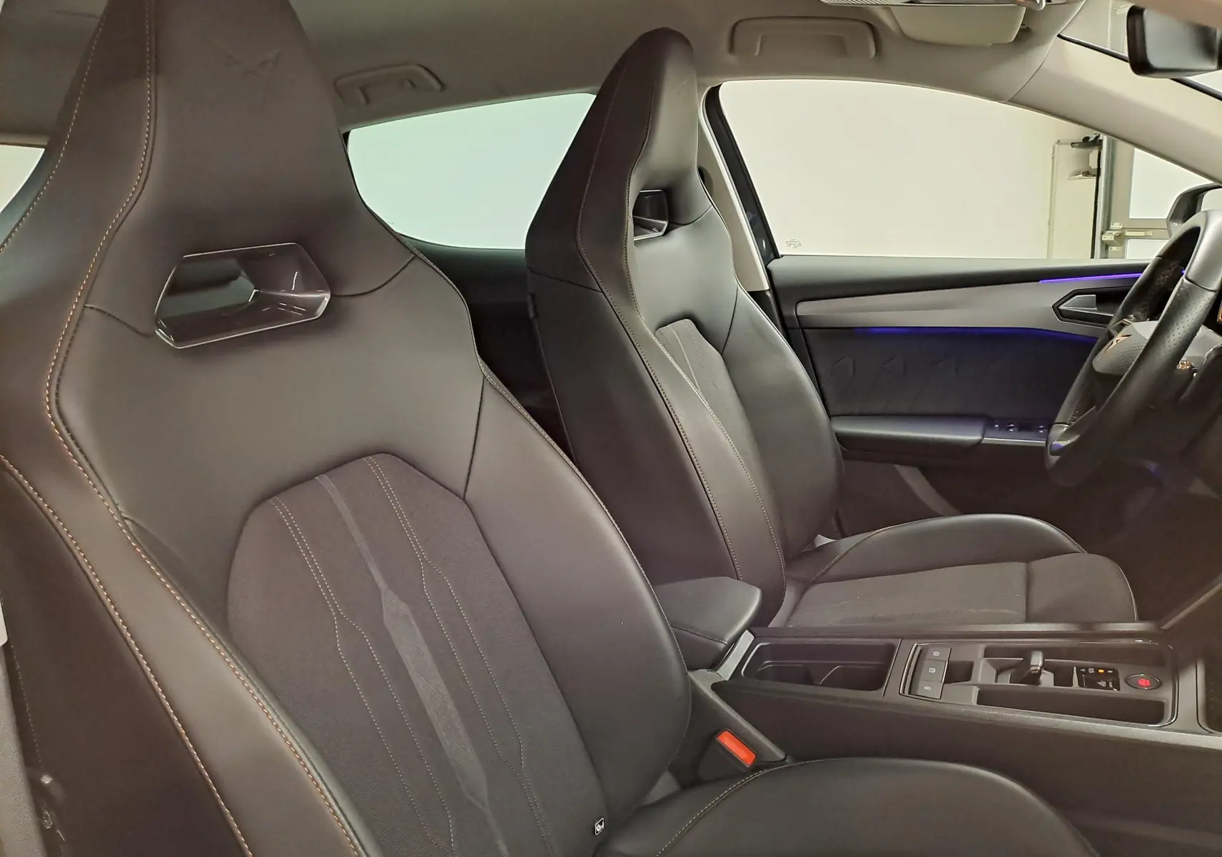 Vue intérieure côté conducteur de la CUPRA Leon 1.5 TSI 150 DS7 V 2023 avec sièges sport noirs et éclairage d'ambiance bleu.