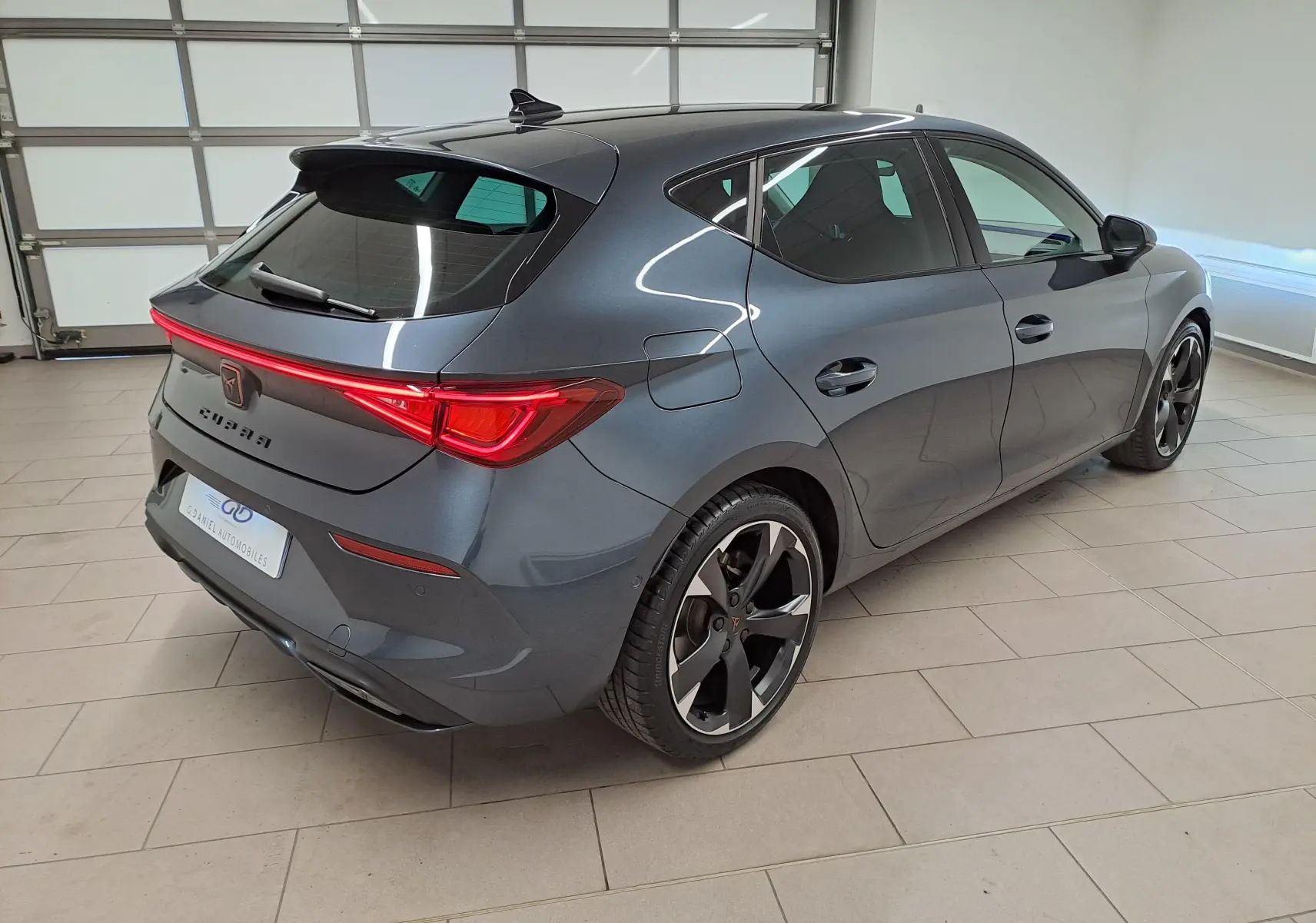 Vue 3/4 arrière droite d'une CUPRA Leon 1.5 TSI 150 DS7 V gris magnétique avec feux arrière LED allumés en intérieur.