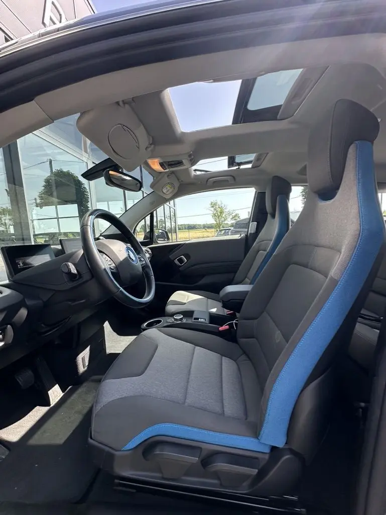 Vue intérieure côté conducteur de la BMW i3 2022 noire, sièges gris avec surpiqûres bleues et toit ouvrant panoramique.