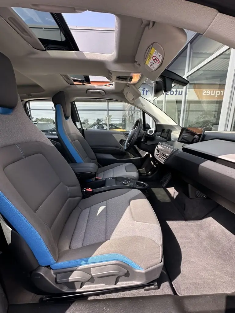 Vue intérieure côté conducteur de la BMW i3 noire 2022, sièges gris avec surpiqûres bleues et toit ouvrant panoramique.