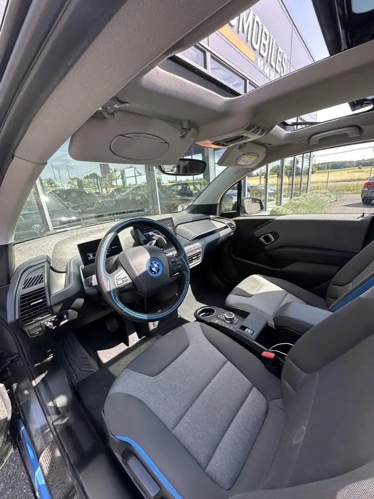 Intérieur de la BMW I3 2022 vue côté conducteur, sièges gris avec surpiqûres bleues et toit ouvrant panoramique.