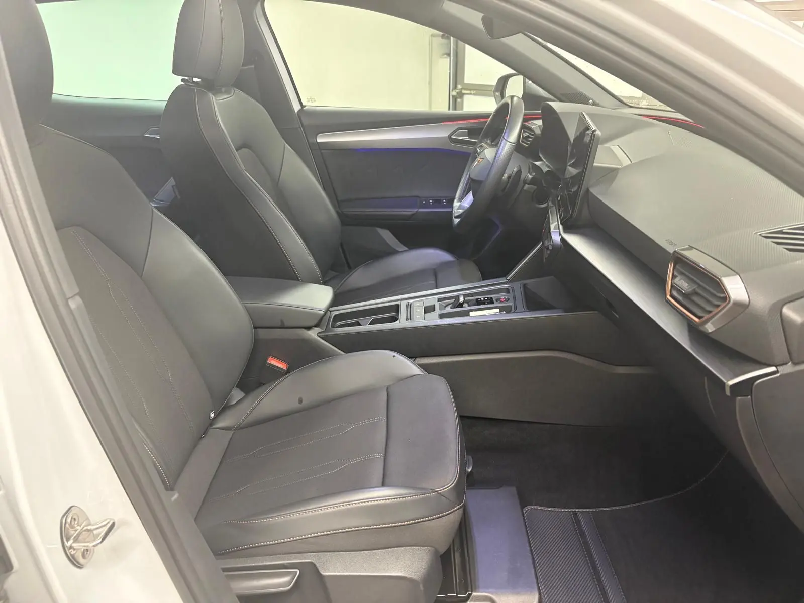 Vue intérieure côté conducteur de la CUPRA Leon 2024 blanche Nevada, avec sièges noirs et tableau de bord moderne.