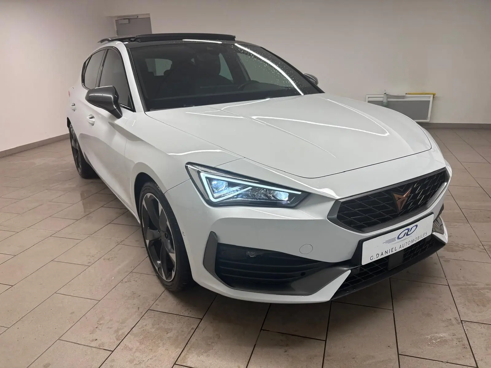 Vue 3/4 avant droite d'une CUPRA Leon 2024 blanc Nevada avec toit ouvrant panoramique et feux LED allumés.
