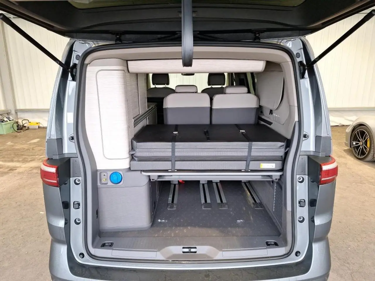 Vue arrière du Volkswagen California T7 gris Indium avec hayon ouvert montrant l'intérieur modulable et la couchette repliée.