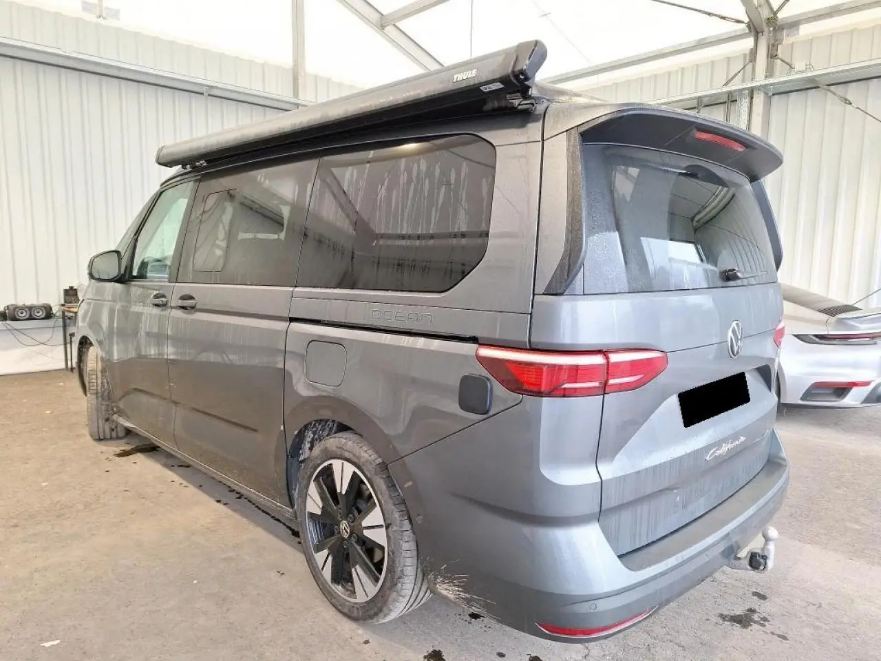 Vue 3/4 arrière droite du Volkswagen California T7 gris Indium avec attelage rabattable et jantes noires brillantes.