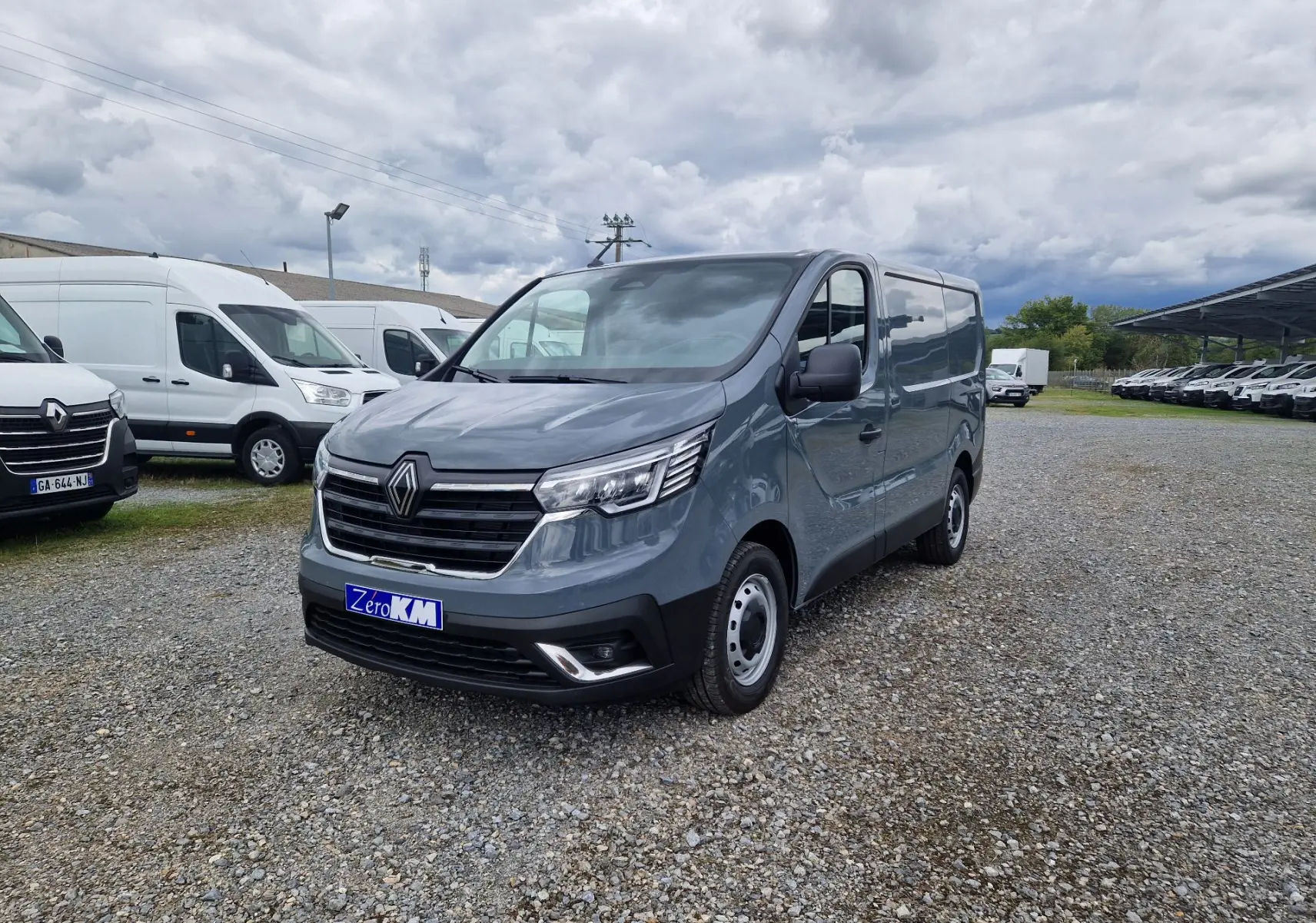 Renault Trafic Fourgon gris urbain vu en 3/4 avant droit sur un parking avec plusieurs utilitaires blancs en arrière-plan.