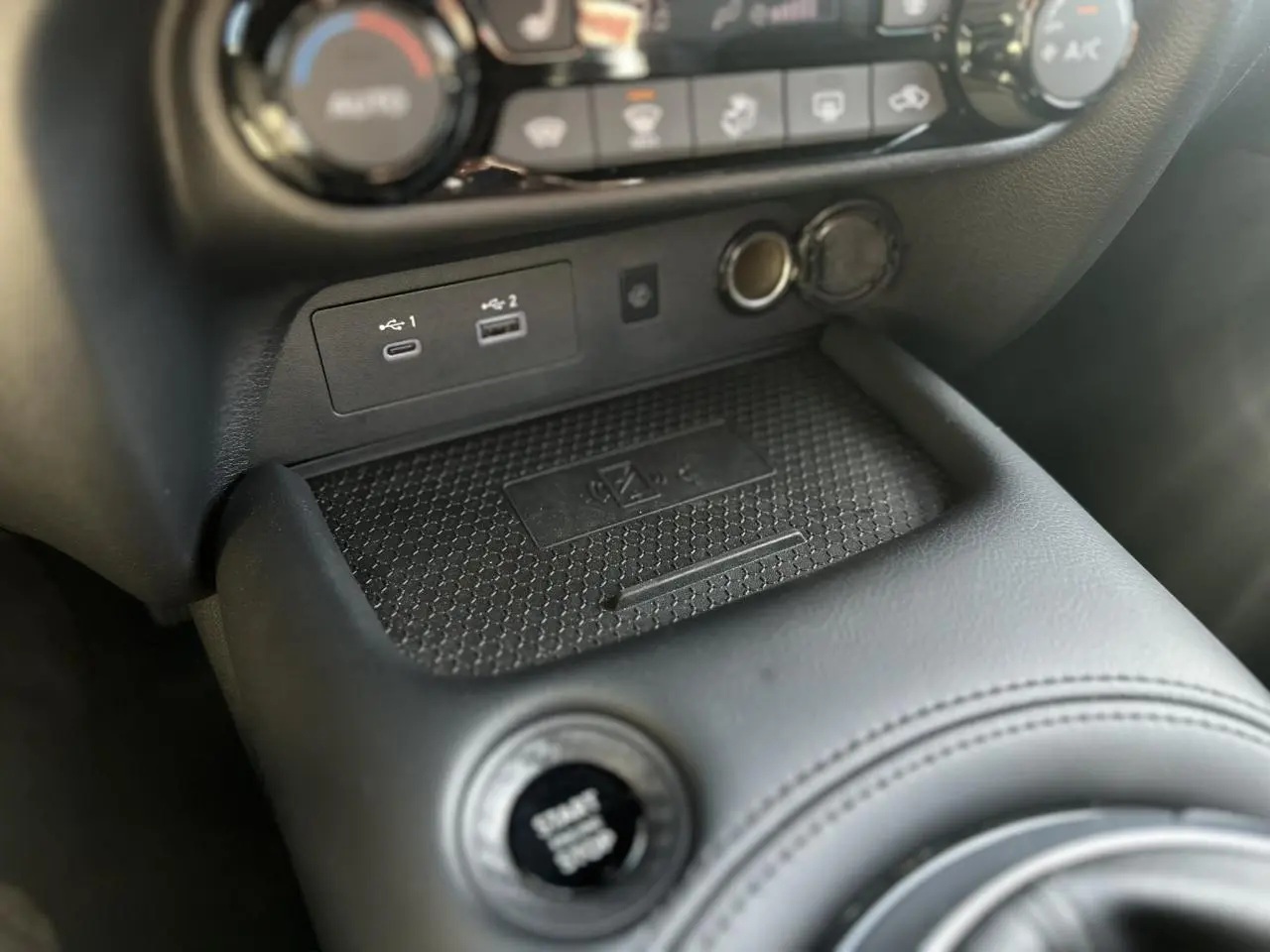 Gros plan sur la console centrale noire du Nissan Juke 2025, montrant le bouton Start/Stop et les ports USB avec chargeur sans fil.