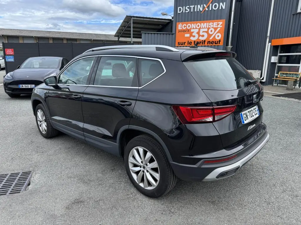 Vue 3/4 arrière droite d'un SUV SEAT Ateca noir métallisé garé devant un bâtiment avec panneaux publicitaires.