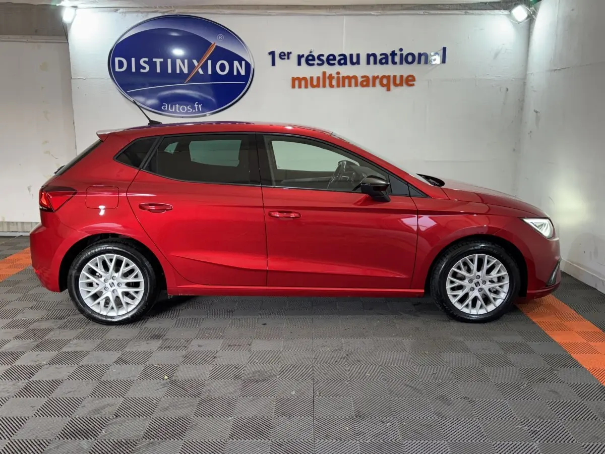 Profil latéral droit d'une SEAT Ibiza 1.0 TSI 110 FR rouge 2024, stationnée en intérieur sur sol damier noir et orange.