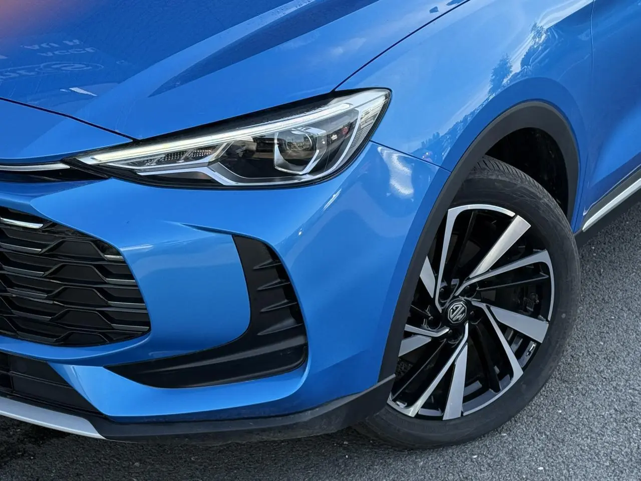 Gros plan sur l'avant droit bleu Como du MG ZS 1.5 Hybrid+ 197 Luxury, mettant en valeur le phare LED et la jante alliage 18 pouces.