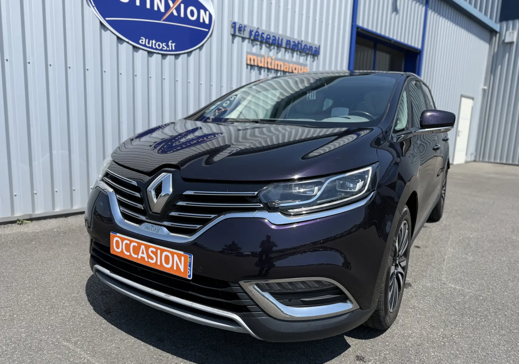 Vue 3/4 avant droite d'un Renault Espace V noir avec calandre chromée et plaque "OCCASION" orange devant un bâtiment industriel.